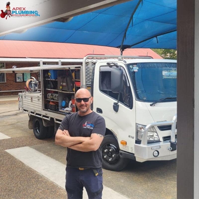 A plumber standing confidently in front of his well-equipped truck under a blue canopy, with a building in the background.
